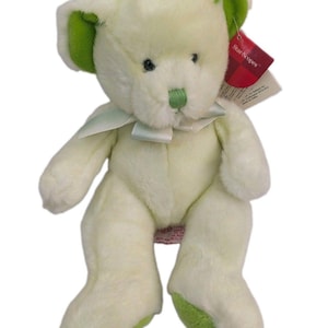 May include: A plush teddy bear with pale yellow fur, featuring bright green accents on the inner ears, nose, and foot pads. The bear has a light-colored ribbon tied around its neck and a red Star-Scopes tag attached to its left ear.