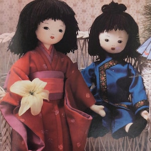 May include: Two handmade cloth dolls with black hair and painted faces. One doll wears a red kimono with a white flower, the other a blue outfit with gold trim. They are seated on a wicker chair.