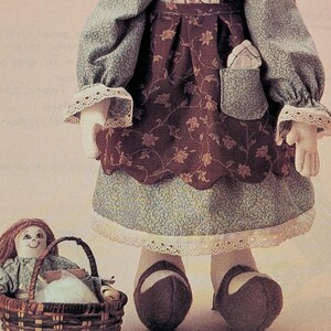 K&ouml;nnte beinhalten: Handgefertigte Stoffpuppe in einem blau-braunen Kleid mit Blumenmuster und wei&szlig;er Spitzenborte. Die Puppe tr&auml;gt braune Schuhe und einen kleinen Korb mit einer kleineren Puppe. Das Kleid hat lange &Auml;rmel und eine Tasche.