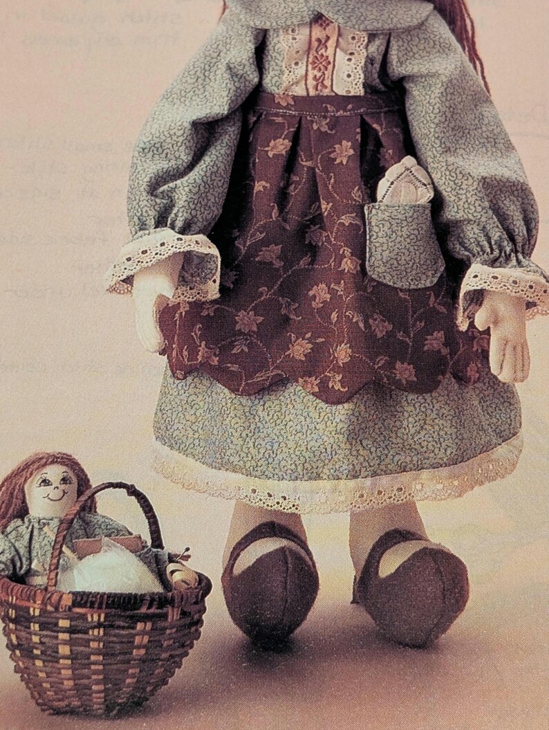 K&ouml;nnte beinhalten: Handgefertigte Stoffpuppe in einem blau-braunen Kleid mit Blumenmuster und wei&szlig;er Spitzenborte. Die Puppe tr&auml;gt braune Schuhe und einen kleinen Korb mit einer kleineren Puppe. Das Kleid hat lange &Auml;rmel und eine Tasche.