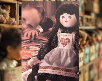 May include: A handmade doll with black hair and a heart-shaped patch on its apron. The doll is wearing a floral print dress and a white apron with red trim. A child is visible in the background.