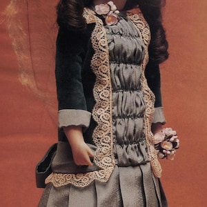 Puede incluir: Una muñeca de porcelana vintage con una chaqueta de terciopelo verde oscuro con ribete de encaje, un vestido gris y un sombrero a juego. La muñeca tiene pelo largo y oscuro y sostiene un pequeño ramo de flores. La muñeca está sobre una base blanca.