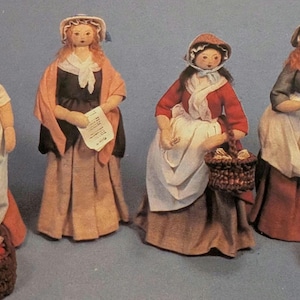 Puede incluir: Cuatro muñecas de tela hechas a mano vestidas con trajes históricos. Cada muñeca lleva un gorro o sombrero, una falda larga y un delantal. Algunas muñecas llevan cestas llenas de productos, mientras que otra sostiene un papel.