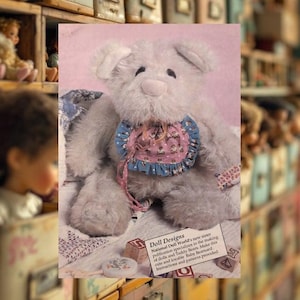 May include: A plush, light gray teddy bear with floppy ears and a pink and blue bib. The bib is trimmed with blue ruffles and has a pink bow. The image includes the text "Doll Designs" and instructions for making dolls and teddy bears.