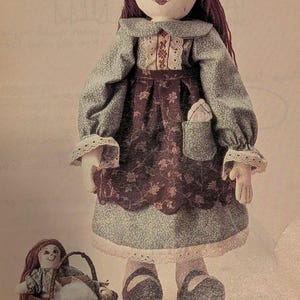K&ouml;nnte beinhalten: Eine handgefertigte Stoffpuppe mit braunem Garnhaar, die ein blaues und braunes Kleid mit Blumenmuster und einer Tasche tr&auml;gt. Eine kleinere Puppe sitzt in einem Weidenkorb. Die Puppe hat ein gemaltes Gesicht und tr&auml;gt eine Haube.