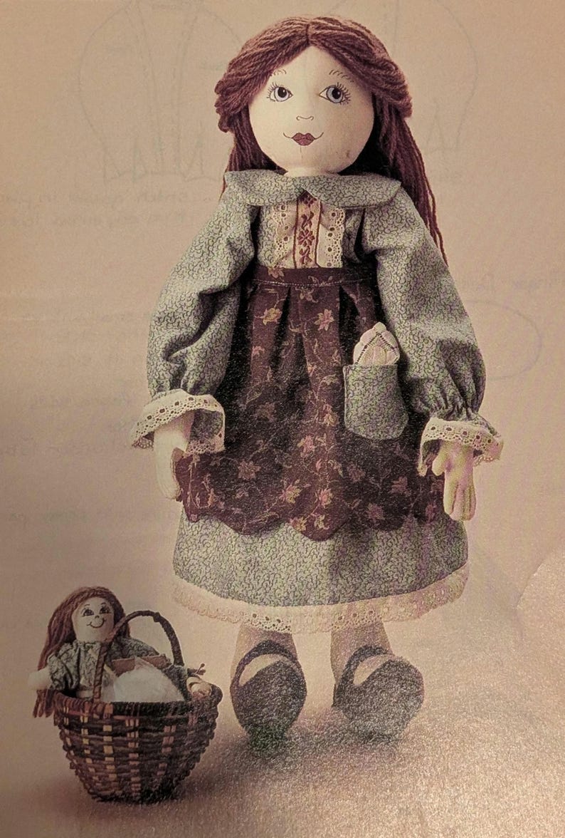 K&ouml;nnte beinhalten: Eine handgefertigte Stoffpuppe mit braunem Garnhaar, die ein blaues und braunes Kleid mit Blumenmuster und einer Tasche tr&auml;gt. Eine kleinere Puppe sitzt in einem Weidenkorb. Die Puppe hat ein gemaltes Gesicht und tr&auml;gt eine Haube.