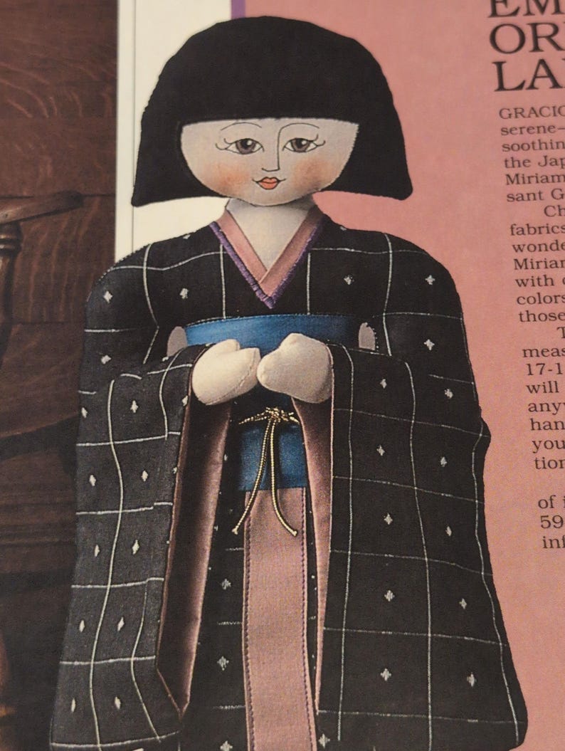 Puede incluir: Mu&ntilde;eca hecha a mano con un kimono negro con un patr&oacute;n de cuadr&iacute;cula blanca y peque&ntilde;os diamantes blancos. El kimono tiene un forro rosa y azul, y una faja azul. La mu&ntilde;eca tiene un peinado bob negro y una cara pintada.