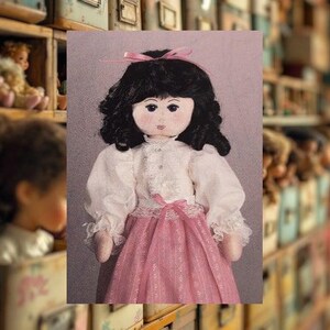 Könnte beinhalten: Handgefertigte Puppe mit schwarzen Locken, einer rosa Schleife und einer weißen Bluse mit Spitzenbesatz. Die Puppe hat einen rosa Rock. Der unscharfe Hintergrund zeigt Vintage-Aufbewahrungsboxen, was auf ein Vintage- oder handwerkliches Thema hindeutet.