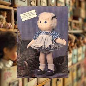 Może przedstawiać: Ręcznie robiona lalka o imieniu "Jody" w niebiesko-białej sukience w paski, białych skarpetkach i niebieskich butach. Lalka ma białe włosy i niebieskie oczy. Obraz zawiera tekst "Jody" zaprojektowany przez Carolee dla czytelników National Doll World.