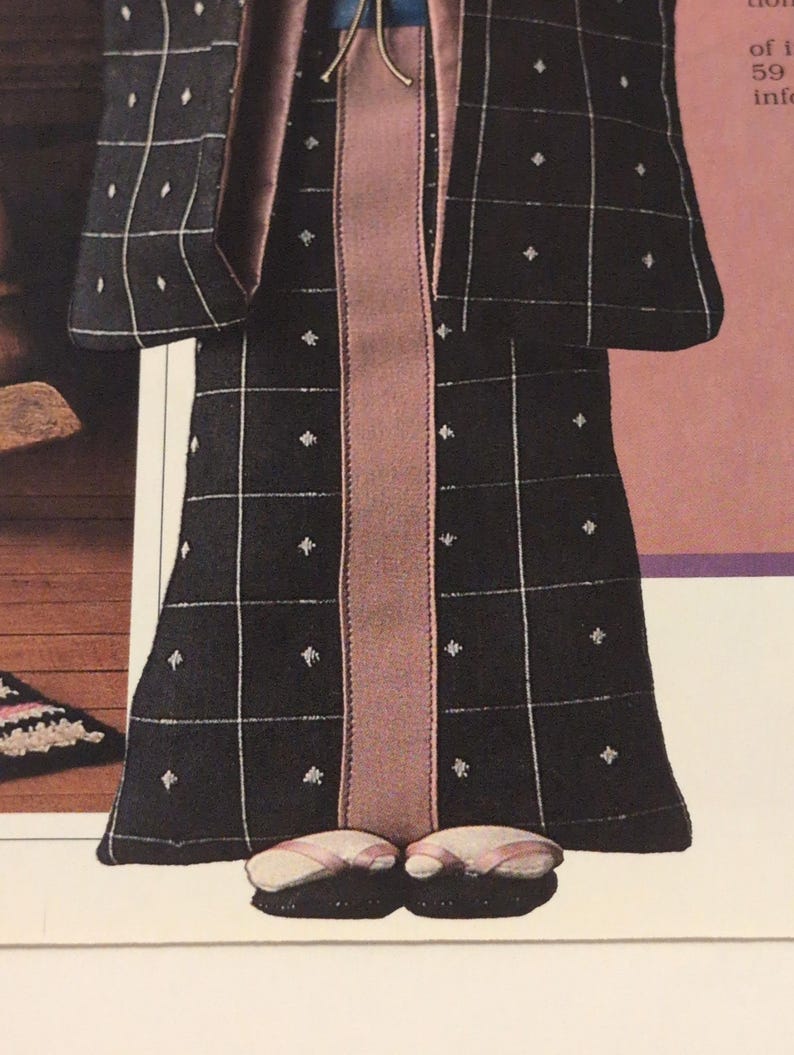 Puede incluir: Mu&ntilde;eca con un kimono negro con un patr&oacute;n de cuadr&iacute;cula blanca y peque&ntilde;os acentos de diamantes blancos. La t&uacute;nica tiene un panel central de color malva y se combina con zapatillas a juego. El atuendo de la mu&ntilde;eca recuerda la vestimenta tradicional japonesa.