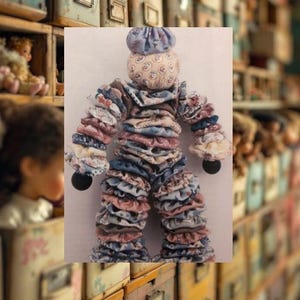 May include: A handmade fabric doll with a layered ruffle design in shades of blue, pink, and cream. The doll has a round head and a small, round black bead for each hand. The doll is standing in front of a wall of vintage drawers.