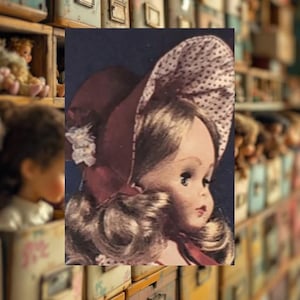 May include: A vintage doll with blonde hair and blue eyes, wearing a burgundy bonnet with white polka dots and a matching ribbon. The doll has a delicate painted face and is part of a collection.