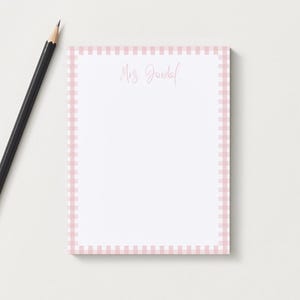 May include: A white notepad with a pink gingham border and the name "Mrs. Gundal" in cursive script. A black pencil with a gold ferrule rests on the white surface next to the notepad. Stationery.