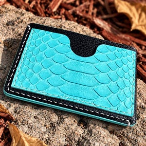 May include: A rectangular card holder featuring a bright turquoise snake-skin textured panel on the front, framed by black leather edges. The design includes a central semi-circular cutout at the top and is held together by visible white stitching.