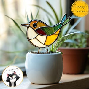 Puede incluir: Adorno de pájaro de vidriera con paneles amarillos, verdes, rojos y blancos. El pájaro está posado en una pequeña maceta de cerámica blanca. La imagen incluye una pegatina "Hobby License".