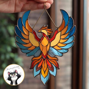 Peut inclure: Un vitrail représentant un phénix aux ailes déployées, avec des couleurs vives de bleu, orange, jaune et rouge. L'œuvre est suspendue par une chaîne dorée. L'image comprend un logo circulaire avec un chat noir et blanc et le texte "Cherishing Glass Co."