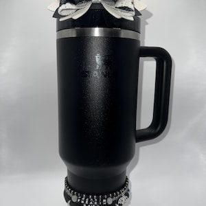 May include: A black Stanley travel mug with a handle, topped with a black and white bow. A stack of bracelets with the text "Freakin Bats" is around the base of the mug. The mug has a silver rim and the Stanley logo.