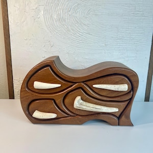Puede incluir: Una caja de joyería de madera con un diseño único y abstracto. La caja tiene tres compartimentos con acentos blancos que parecen hueso. La caja está hecha de madera oscura y tiene un acabado liso y pulido.