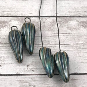 Op de afbeelding: Vier blauwgroene keramische hangerkralen met verticale ribbels en een metallic glans. Elke kraal is druppelvormig en heeft een kleine lus aan de bovenkant om te rijgen. De kralen worden gepresenteerd tegen een lichte houtnerf achtergrond.