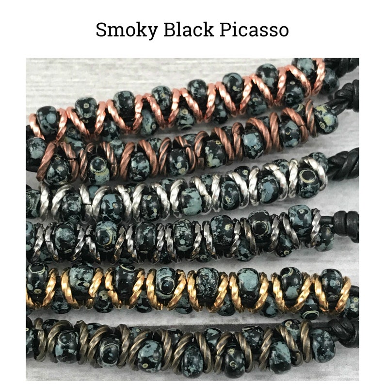 May include: A close-up of a multi-strand bracelet with black and white beads. The beads are interspersed with copper, silver, and gold wire. The bracelet is made with a variety of materials and has a unique, handcrafted look.