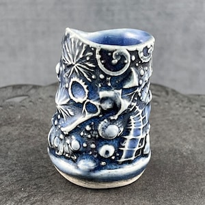 May include: A small, blue ceramic cup with a white glaze and a textured design. The cup has a unique, abstract pattern that resembles stars, swirls, and bubbles.