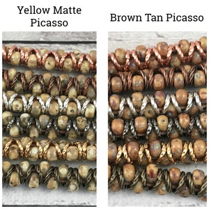 May include: Close-up of two strands of beads, one in a yellow matte Picasso color and the other in a brown tan Picasso color. Both strands feature a mix of copper, silver, and gold wire accents.