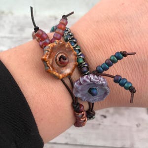 May include: Three bracelets with brown leather cord and colorful beads. One bracelet has a brown ceramic flower charm, another has a purple ceramic flower charm, and the third has a silver metal chain.