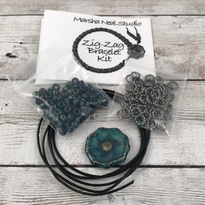 May include: A kit for making a zig-zag bracelet. The kit includes blue and green beads, silver jump rings, black cord, and a teal ceramic charm. The kit is packaged in a clear plastic bag with a white card that says "Marsha Neal Studio" and "Zig-Zag Bracelet Kit".