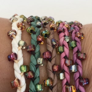 May include: Five braided bracelets with iridescent beads. The bracelets are in shades of white, green, brown, pink, and purple.