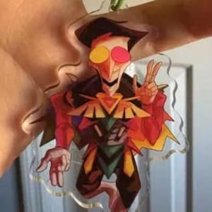 May include: A clear acrylic keychain featuring a colorful cartoon character with a red and black outfit, yellow and pink eyes, and a peace sign gesture. The keychain has a green metal ring and clasp.