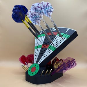 Könnte beinhalten: Ein schwarzer Dart-Halter in Form einer Dartscheibe mit grünem und rotem Ziel-Design. Der Halter zeigt mehrere Darts mit blauen und weißen Flights. Zusätzliche Darts mit roten und lila Flights befinden sich in der Basis.