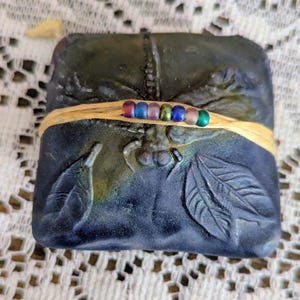 May include: A dark blue, rectangular soap bar with embossed leaf and insect designs. The soap is wrapped with a tan ribbon adorned with colorful beads. The soap has a textured appearance and a slight iridescent sheen.