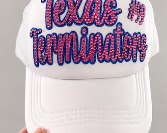May include: White trucker hat with the text "Texas Terminator" in pink rhinestones outlined in blue. The hat has a white mesh back and a curved brim. A small pink rhinestone image of a longhorn is on the right side.