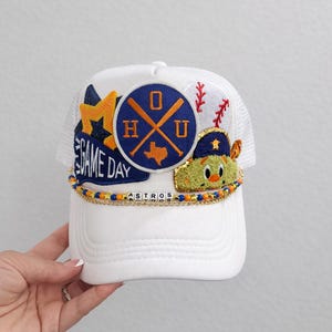 May include: White baseball cap with mesh back and embroidered patches. Patches include a baseball, a cartoon character, and a logo with the letters "H" and "U" and the Texas outline. The text "GAME DAY" is also visible.
