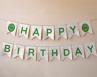 May include: A banner with the words "HAPPY BIRTHDAY" in green letters. The banner is made of white pennants with a floral pattern and turtle shell graphics. The banner is strung on a thin white string.