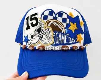 May include: A white and blue trucker hat featuring a football-themed design with the number 15, a football helmet, a brown football, a blue pennant with the text GAME DAY, gold sequin stars, and a beaded trim with the name ELWELL.