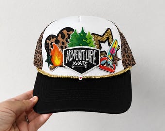 May include: A trucker hat with a white front panel, black brim, and leopard print mesh back. The front features a central patch reading ADVENTURE AWAITS, surrounded by embroidered patches of a campfire, pine trees, a gold star, and a surfboard.