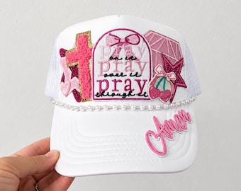 May include: A white trucker hat featuring a central patch with the text 'pray on it pray over it pray through it' and a pink bow. Additional patches include a pink cross, a diamond, a star, and cherries. The hat is accented with a pearl trim and the word 'Amen'.