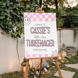 May include: A wooden easel holds a white sign with pink checkered borders and text. The sign reads "welcome to Cassie's little miss THREENAGER birthday party!" with a smiley face and star graphics. The sign is outdoors with greenery in the background.