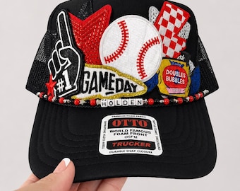 May include: A black trucker hat featuring a foam front panel with multiple patches including a baseball, a red star, a number one foam finger, and a red and white checkered flag. The hat includes a beaded trim with the name HOLDEN and an OTTO label.