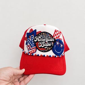 May include: A red and white trucker hat featuring a central circular patch with the text American Babe, surrounded by various embroidered patches including a peace sign, a smiley face, and stars in red, white, and blue with beaded trim.