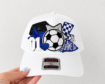 May include: A white baseball cap featuring a central soccer ball patch, a blue glitter star, a black geometric shape, a blue heart, the number 01, a blue and white checkered flag, a silver star, and a blue patch with the text GAME DAY.