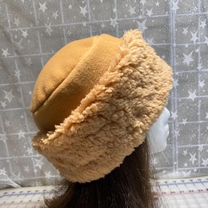 May include: A tan fleece hat with a fluffy, light brown trim. The hat is displayed on a mannequin head with brown hair, showcasing its shape and texture. The hat is designed to keep the wearer warm.