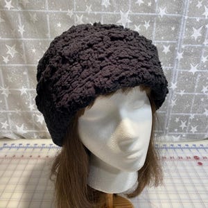 May include: A dark brown, textured crochet hat. The hat is displayed on a white mannequin head with brown hair. The background is a light gray fabric with white star patterns.