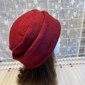 May include: A deep red fleece hat with a rounded crown and a wide, folded brim. The hat is displayed on a mannequin head, positioned next to a white cutting mat with visible measurement markings in inches.
