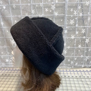 May include: A black, fleece-lined winter hat with a folded brim. The hat is displayed on a mannequin head with brown hair. The hat's texture appears soft and plush, suitable for cold weather. The background features a white star pattern.