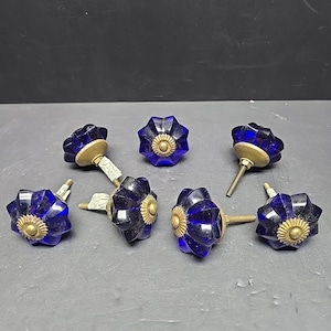 Puede incluir: Un conjunto de siete tiradores de armario de cristal azul cobalto con diseño de estrella. Cada tirador tiene una base y un tornillo de color latón. Los tiradores están dispuestos sobre una superficie gris.