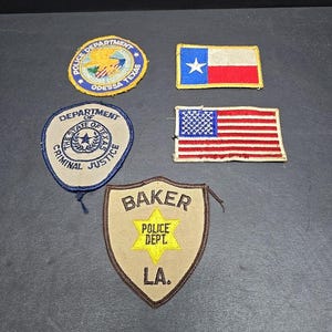 May include: A selection of embroidered patches with law enforcement and patriotic designs. The patches display the Texas state flag, the American flag, and police department emblems, including "Odessa Texas" and "Baker Police Dept. LA."