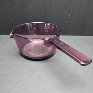 Puede incluir: Una cacerola de vidrio morado vintage con asa y boquilla. La cacerola está hecha de vidrio translúcido de color púrpura oscuro. El asa está unida al costado de la sartén. La cacerola está sobre una superficie gris.