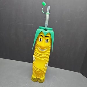Puede incluir: Una taza para beber con forma de maíz con tapa y pelo verdes, una cara sonriente y una pajita transparente. La taza es amarilla con una superficie texturizada y la pajita tiene una tapa verde.